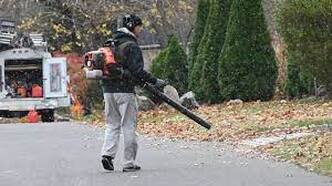 Man using a leaf blower along a roadway with a service truck and pine trees in the background